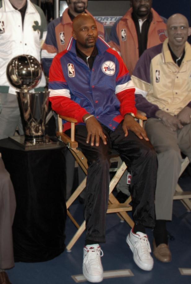 Ein Mann in einer Jacke der Philadelphia 76ers sitzt neben einer Trophäe.