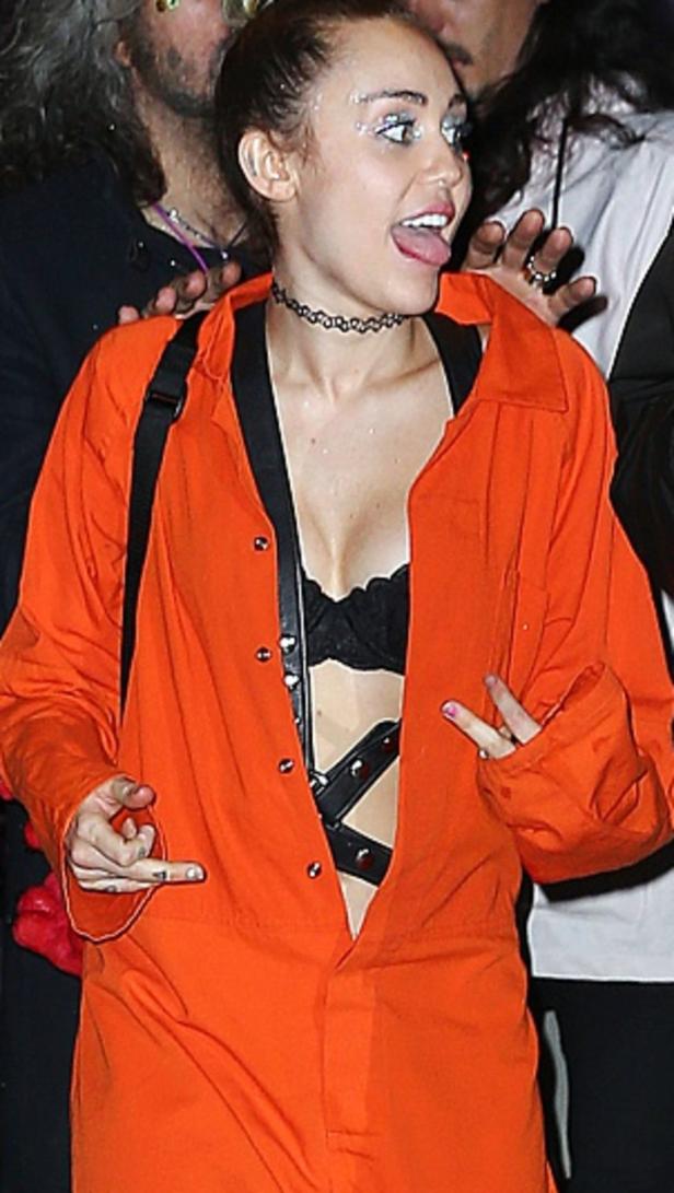 Miley Cyrus in einem orangefarbenen Overall mit Glitzer um die Augen streckt die Zunge heraus.