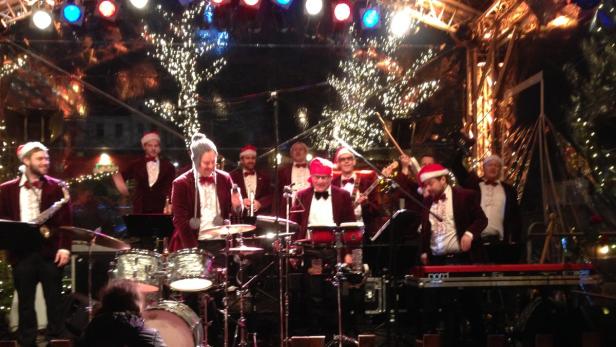 Eine Band mit Weihnachtsmützen spielt auf einer Bühne vor einem Weihnachtsbaum.