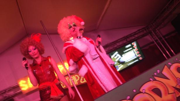 Zwei Dragqueens treten in auffälligen Kostümen und Perücken auf einer Bühne auf.