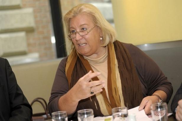 Eine blonde Frau mit Brille gestikuliert an einem Tisch während einer Besprechung.