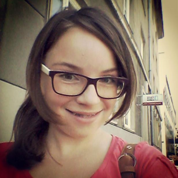 Eine lächelnde junge Frau mit Brille und Zahnspange vor einem Gebäude.