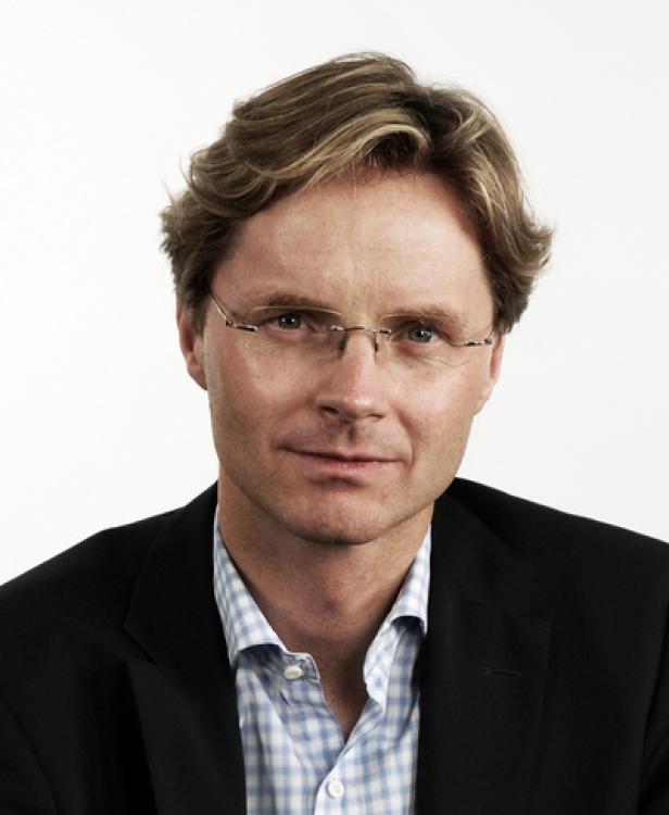 Porträt eines Mannes mit blonden Haaren, Brille und dunklem Anzug vor einem weißen Hintergrund.
