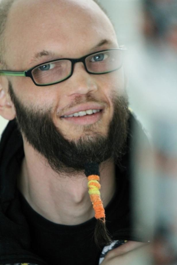 Ein Mann mit Brille und Bart lächelt in die Kamera. Der Bart ist mit bunten Bändern verziert.
