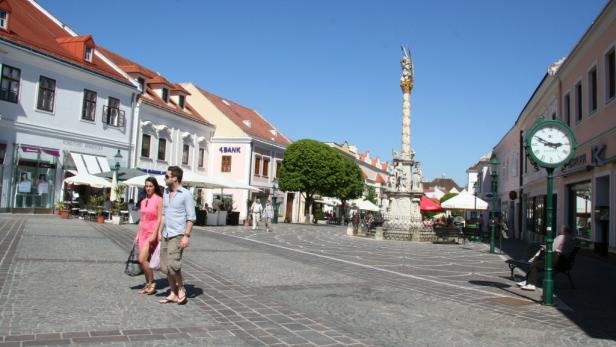 Ein Paar spaziert über einen belebten Platz mit barocker Säule in einer europäischen Stadt.