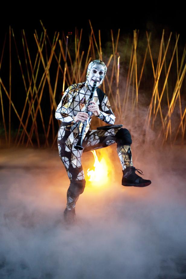 Ein Künstler mit Gesichtsbemalung spielt Klarinette vor einem Feuer und Bambusstangen.