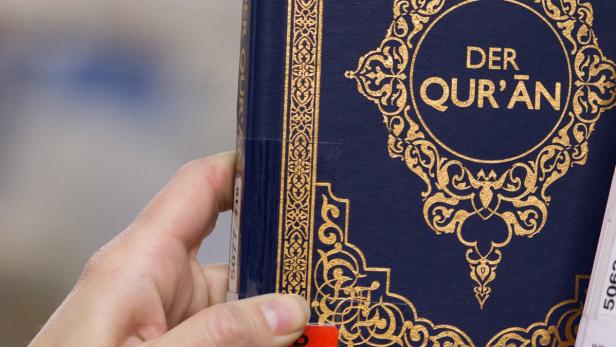 Eine Hand hält ein blaues Buch mit dem Titel „Der Qur’an“ in goldener Schrift.