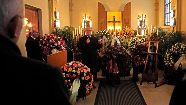 Ein Mann spricht bei einer Trauerfeier vor Kränzen und einem Foto des Verstorbenen.