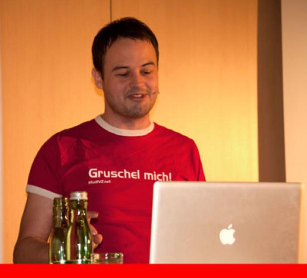 Ein Mann mit einem roten T-Shirt mit der Aufschrift „Gruschel mich!“ steht vor einem Laptop.