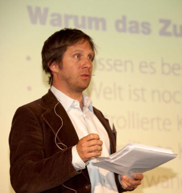 Ein Mann in einem braunen Sakko hält Papiere in der Hand und spricht vor einem Publikum.
