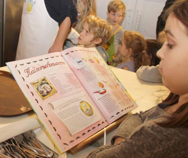 Ein Mädchen liest ein Kochbuch mit dem Rezept für Kaiserschmarrn, während andere Kinder zusehen.