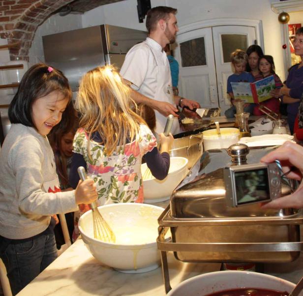 Kinder rühren Teig in Schüsseln, während ein Koch im Hintergrund Pfannkuchen zubereitet.