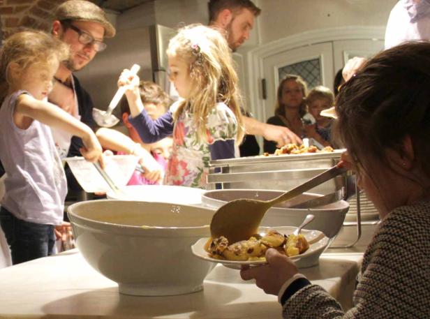 Kinder und Erwachsene schöpfen Essen in einer Küche auf Teller.