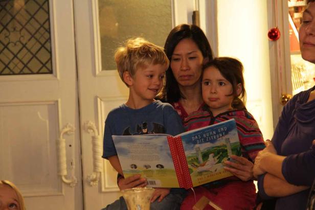 Eine Frau liest mit einem Jungen und einem Mädchen das Buch „Das Huhn und die Zitrone“.