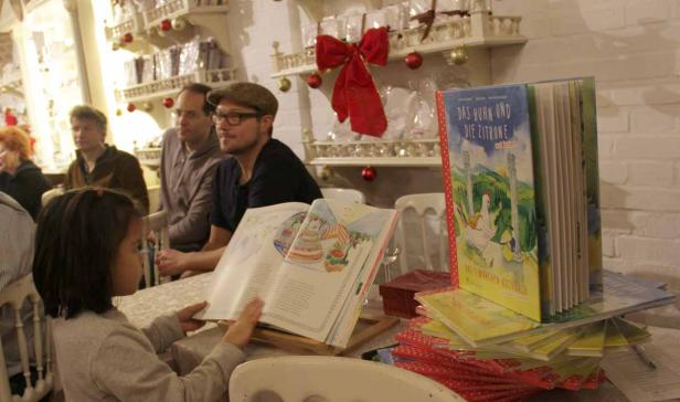 Ein kleines Mädchen betrachtet ein aufgeschlagenes Bilderbuch, während Erwachsene zusehen.