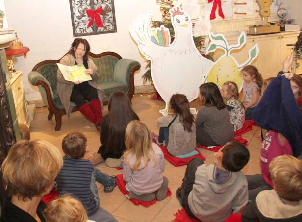 Eine Frau liest einer Gruppe von Kindern eine Geschichte vor, im Hintergrund eine große Hühnerdekoration.