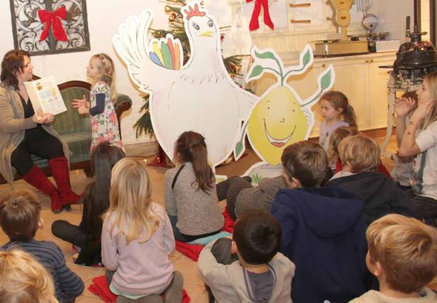 Eine Frau liest einer Gruppe von Kindern eine Geschichte vor, im Hintergrund eine Hühner- und Zitronen-Dekoration.