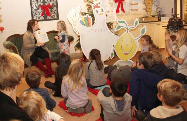 Eine Frau liest einer Gruppe von Kindern eine Geschichte vor, im Hintergrund eine Hühner- und Zitronen-Dekoration.