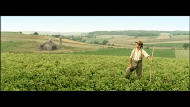 Ein junger Mann mit Hut und Hosenträgern steht auf einem Feld mit einem Stock in der Hand.