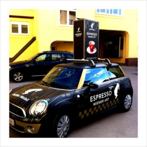 Ein Mini Cooper mit Werbung für „Espresso Wiener Art“ und einem großen Espresso-Werbeaufbau auf dem Dach.
