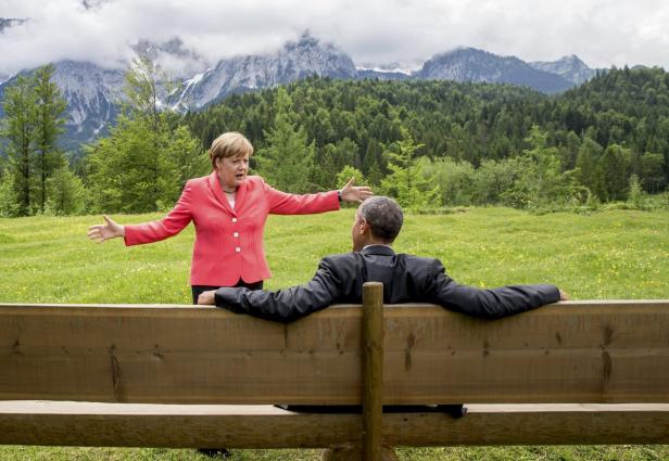 Angela Merkel gestikuliert vor Barack Obama, der auf einer Bank vor einer Bergkulisse sitzt.