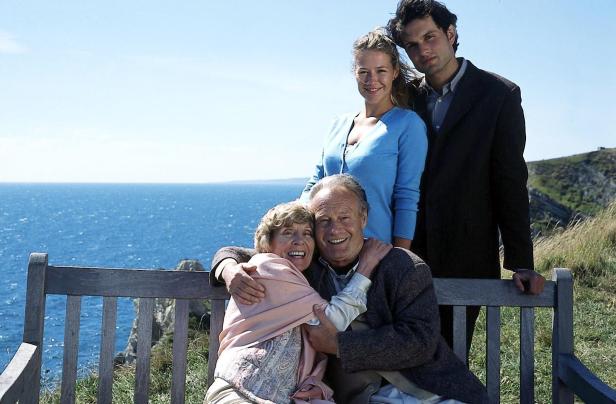 Eine ältere Frau und ein älterer Mann umarmen sich auf einer Bank am Meer, während eine junge Frau und ein junger Mann dahinter stehen.