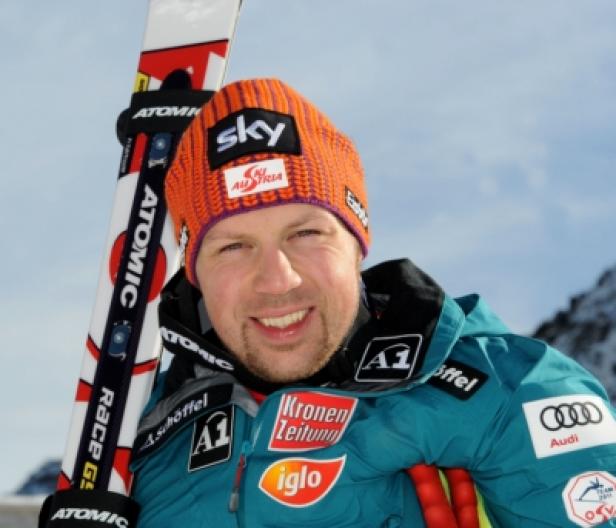 Ein lächelnder Skirennläufer mit Mütze und Skiern vor verschneiten Bergen.