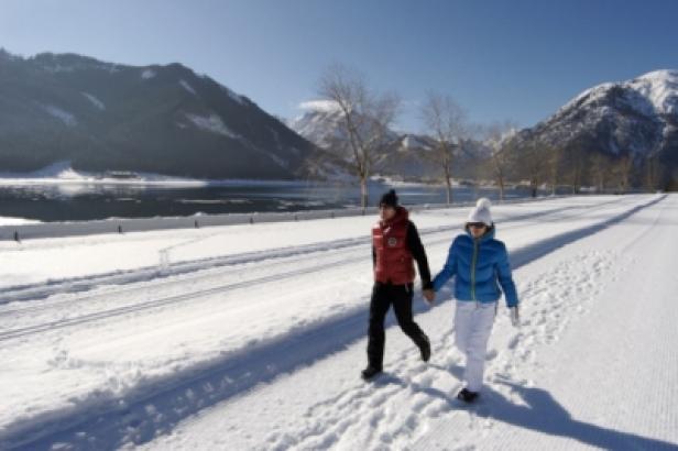Ein Paar geht Hand in Hand auf einem verschneiten Weg entlang eines Sees mit Bergen im Hintergrund.
