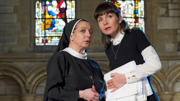 Eine Nonne und eine Frau mit einem Stapel weißer Stoffe stehen vor einem Buntglasfenster.