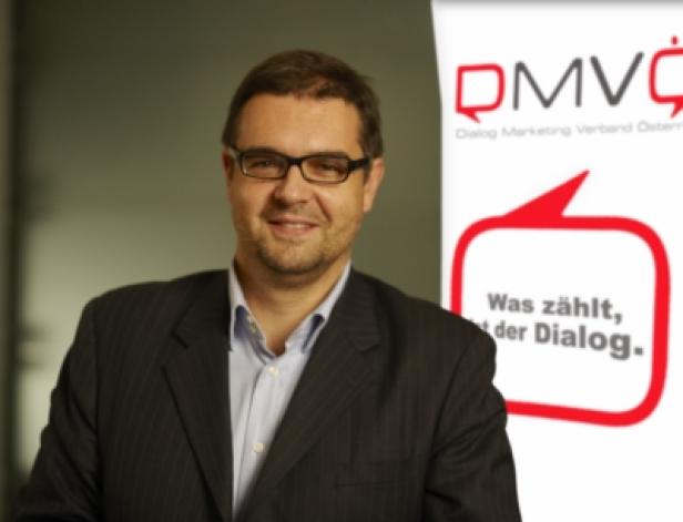 Ein Mann mit Brille und Anzug steht vor einem DMVC-Logo.