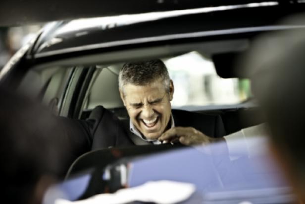 Ein Mann im Anzug sitzt im Auto und lacht.