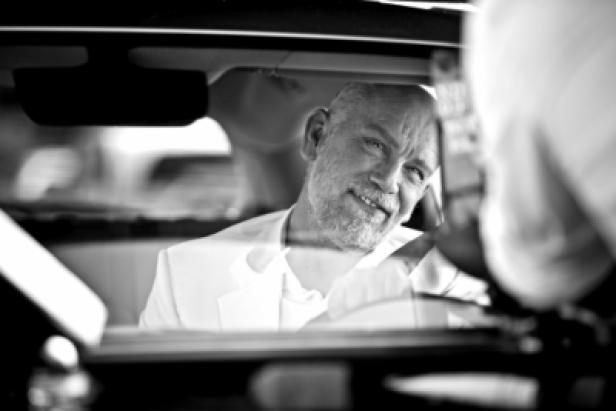 John Malkovich sitzt lächelnd in einem Auto.