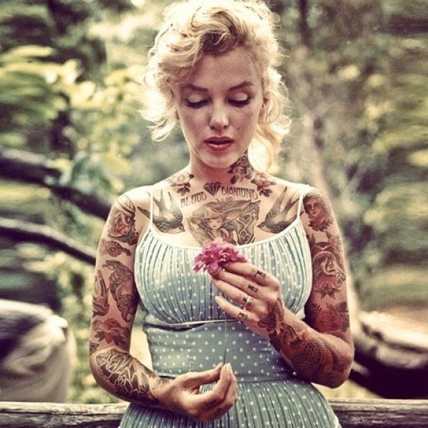 Eine Frau mit blonden Locken und zahlreichen Tattoos betrachtet eine rosa Blume.