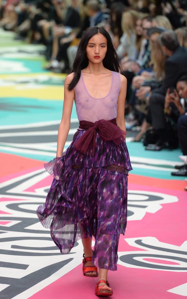 Eine Frau mit dunklen Haaren läuft in einem lila Kleid über einen Laufsteg.