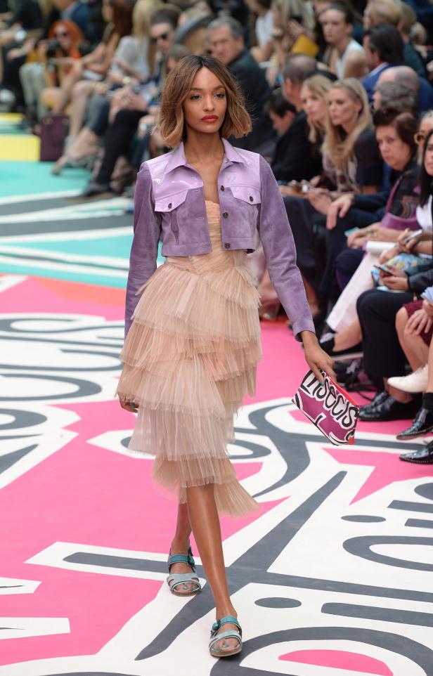 Ein Model präsentiert ein Kleid mit Rüschen und eine lila Jacke auf einem Laufsteg.