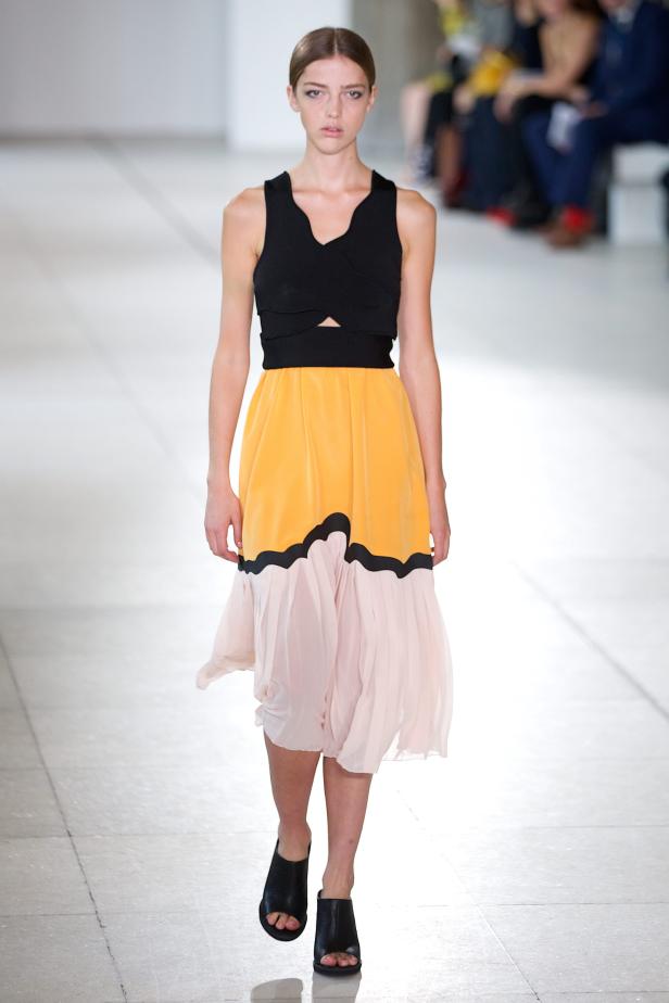Eine Frau präsentiert ein Kleid mit schwarzem Oberteil und gelb-rosa Rock auf einem Laufsteg.