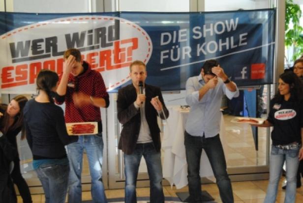Männer stehen auf einer Bühne vor einem Banner mit der Aufschrift „Wer wird esponiert? Die Show für Kohle“.