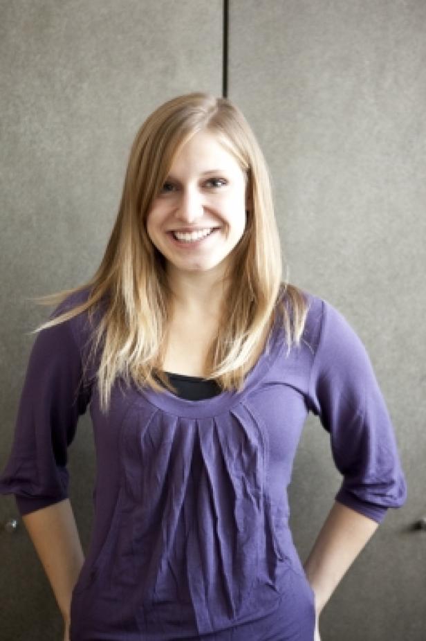 Eine lächelnde Frau mit blonden Haaren und einem lila Oberteil steht vor einer grauen Wand.