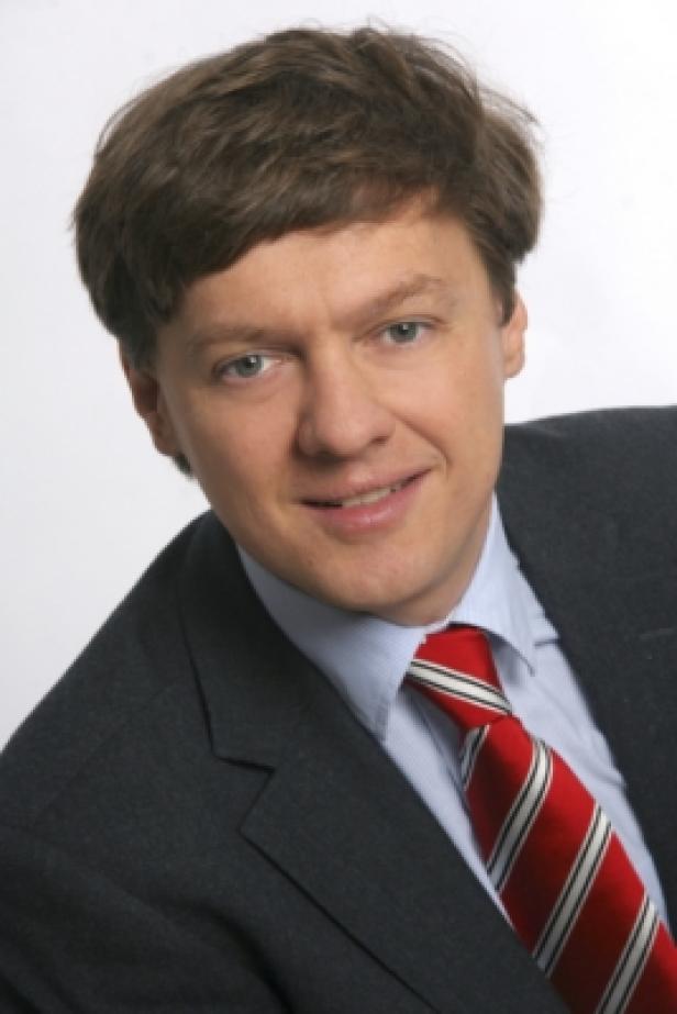 Ein Mann mit braunen Haaren, Anzug und rot-gestreifter Krawatte lächelt.