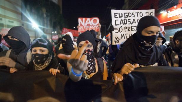 Vermummte Demonstranten protestieren mit Schildern und zeigen den Mittelfinger.