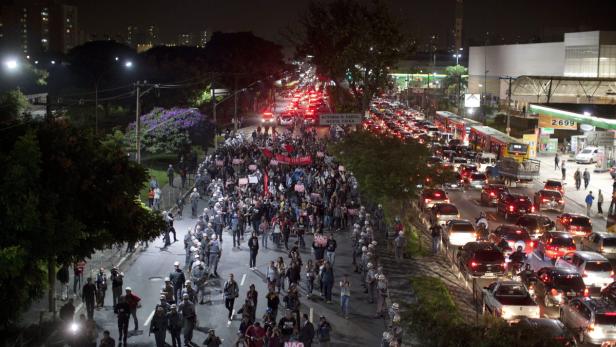 Bei Nacht demonstrieren Menschen auf einer Straße neben einer stark befahrenen Straße.