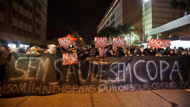 Eine Menschenmenge demonstriert mit einem Banner „Sem Saúde Sem Copa“ und Schildern gegen die Fußball-Weltmeisterschaft.