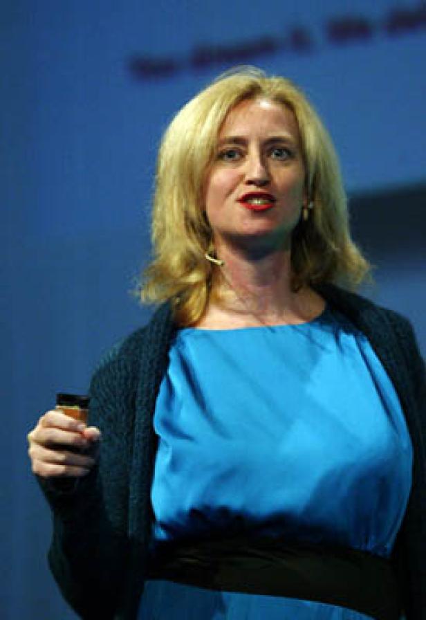 Eine blonde Frau in einem blauen Kleid hält ein Glas und spricht vor einem Publikum.