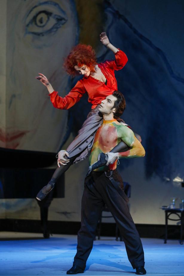 Ein Tänzer trägt eine Tänzerin auf seinen Schultern vor einem gemalten Hintergrund.