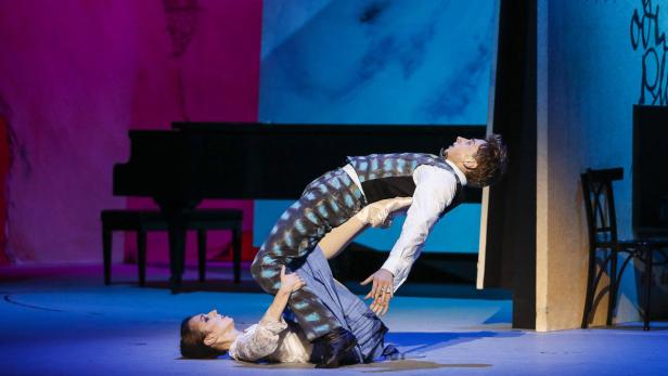 Ein Tänzerpaar führt eine Hebefigur auf einer Bühne vor einem Klavier auf.