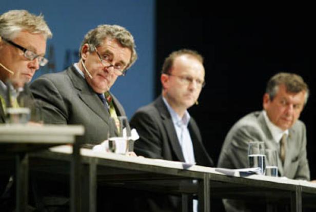 Vier Männer sitzen an einem Tisch während einer Konferenz oder Podiumsdiskussion.
