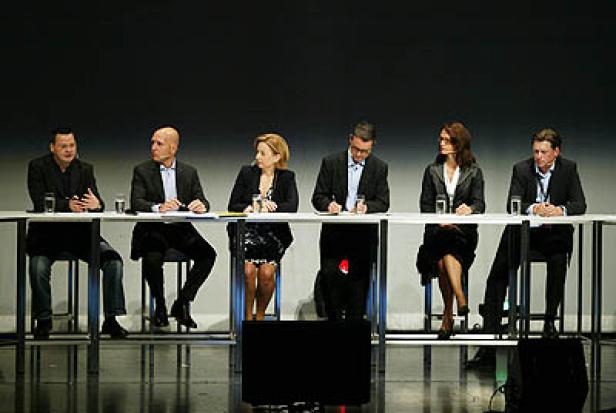 Sechs Personen sitzen an einem Tisch auf einer Bühne.