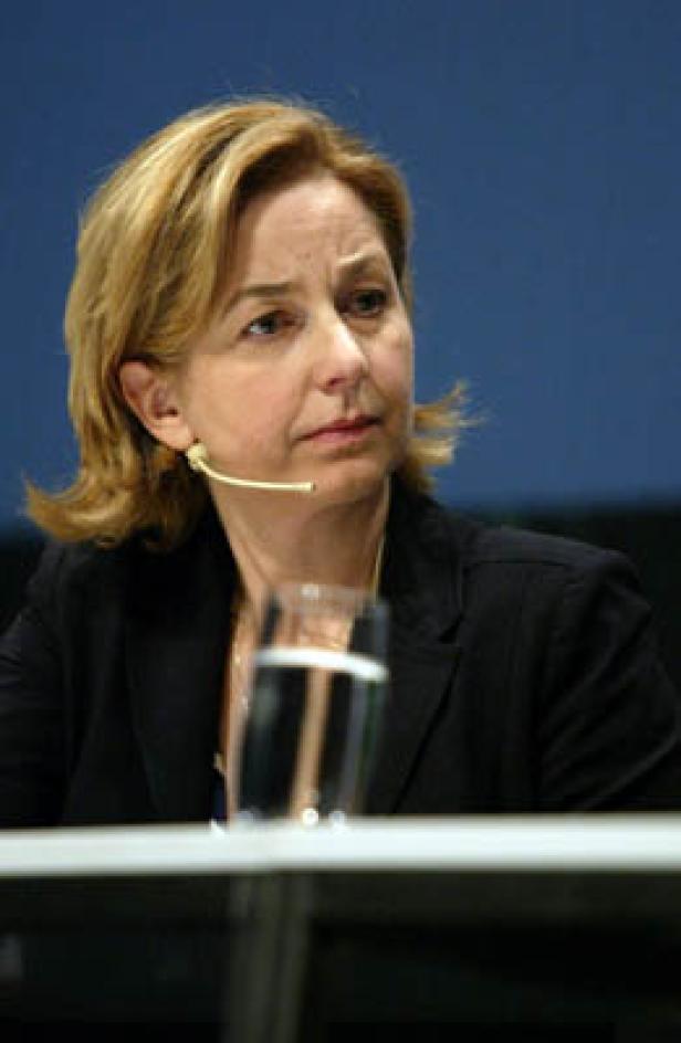 Eine Frau mit blonden Haaren und einem schwarzen Blazer spricht bei einer Konferenz.