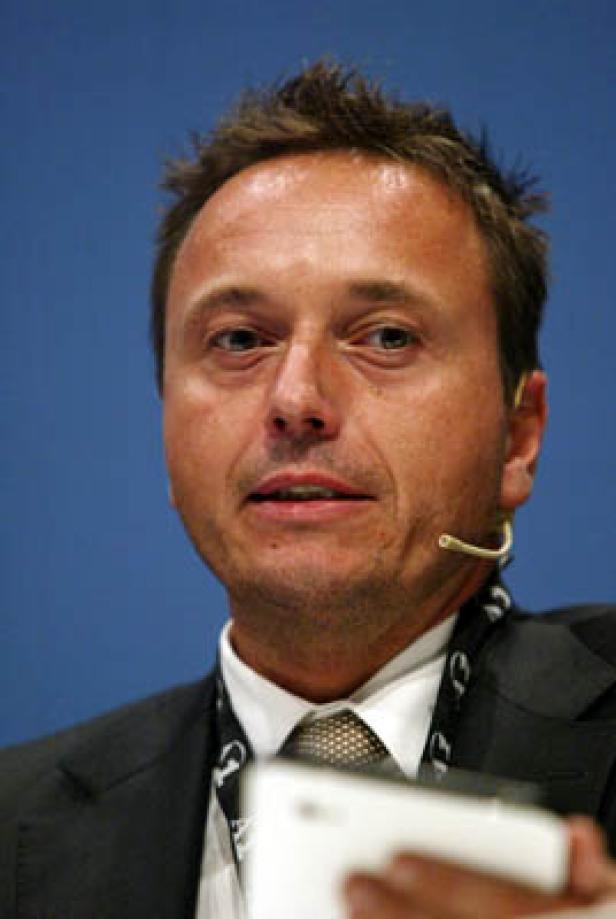 Ein Mann mit Anzug und Mikrofon spricht vor einem blauen Hintergrund.
