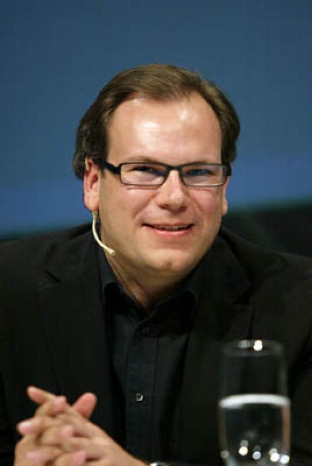 Ein Mann mit Brille und Anzug sitzt an einem Tisch mit einem Mikrofon am Ohr.